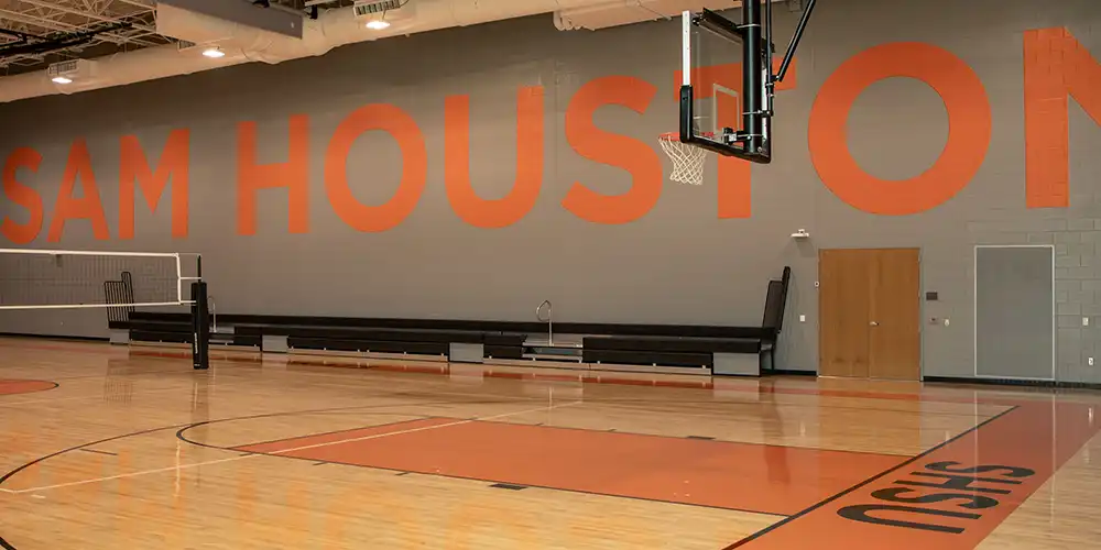 Empty basketball court with orange lettering “SAM HOUSTON” on a gray wall. Volleyball net setup on the left side and basketball goal on the right. Black lettering “SHSU” inside of orange border on the court. 