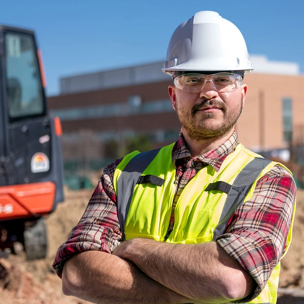 Male construction worker looking forward at you, wearing a bright green safety vest and hard hat, standing inside a construction site.