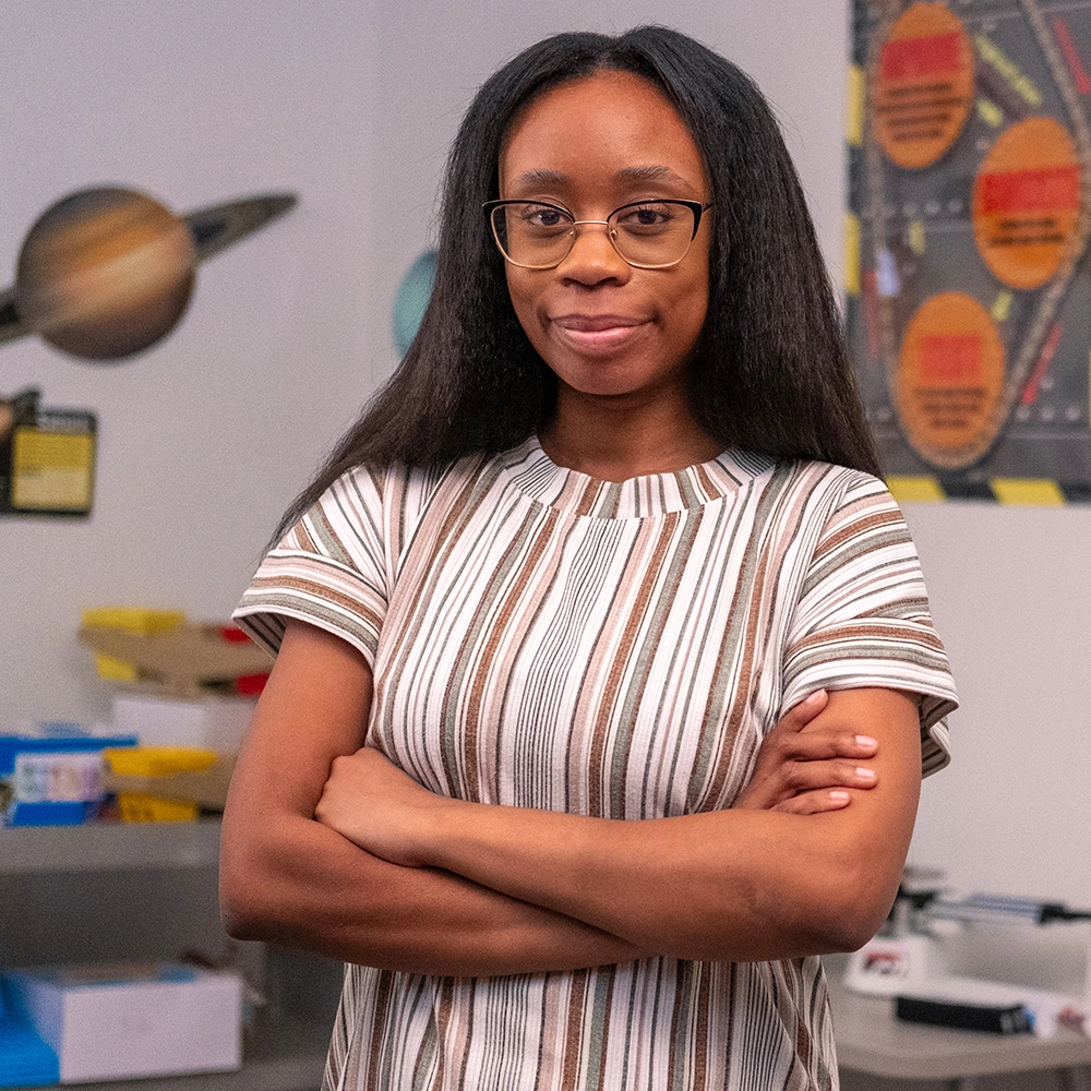 Female science teaching looking forward at you, wearing a striped blouse, and standing within a science classroom