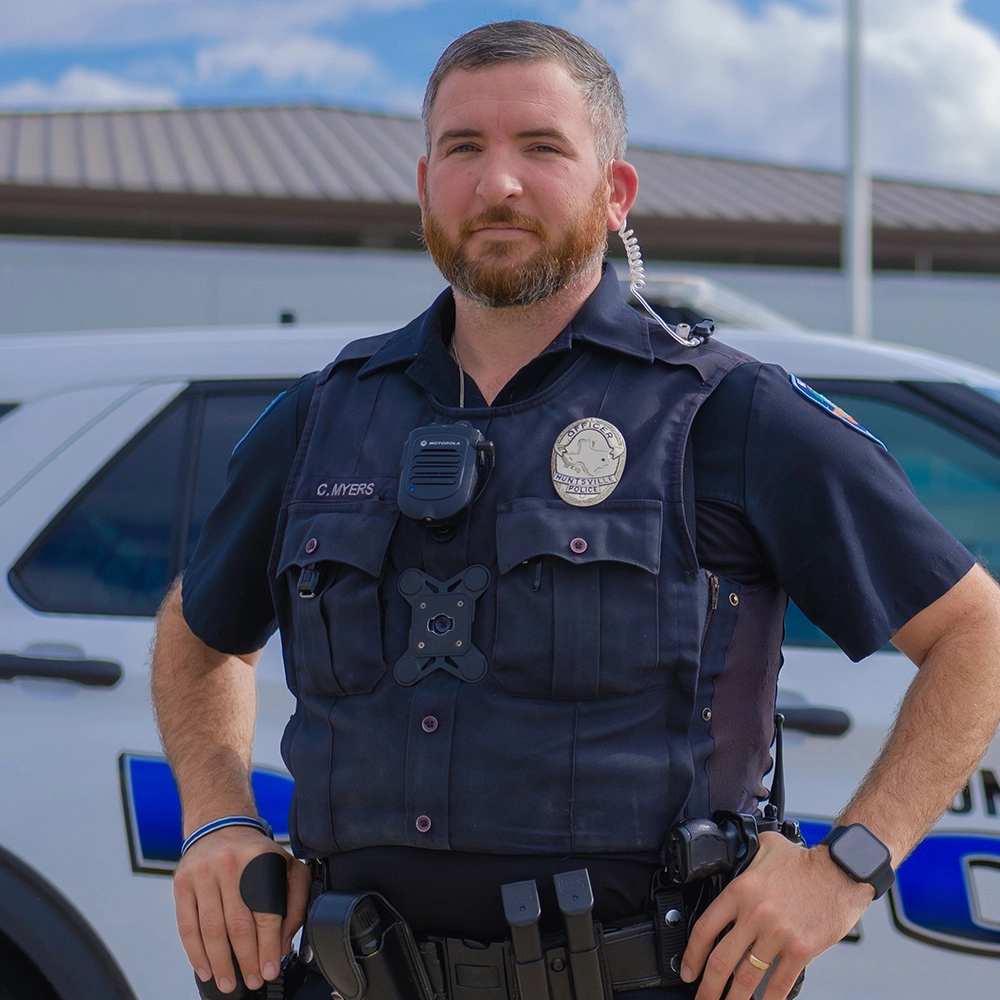 Male police officer looking forward at you, wearing a police uniform, standing in front of their police vehicle (SUV)