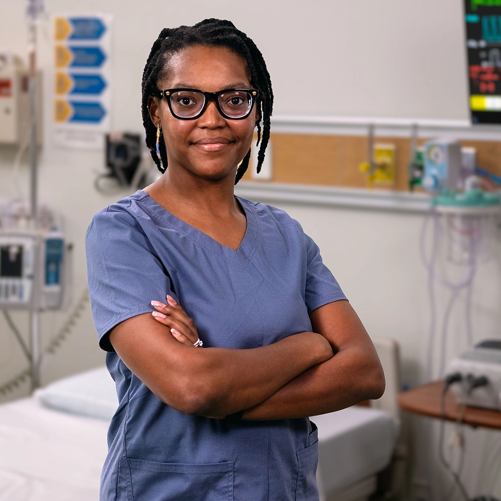 Female nurse looking forward at you, wearing scrubs, standing next to a patient bed in an emergency room.