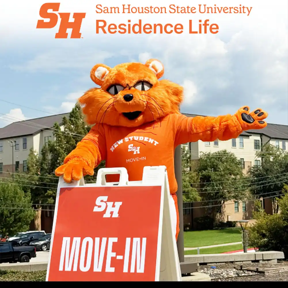 Mascot at Sam Houston State University wearing an orange “New Student Move-In” shirt stands outdoors by residence halls, holding an SH Move-In sign and waving to welcome students.
